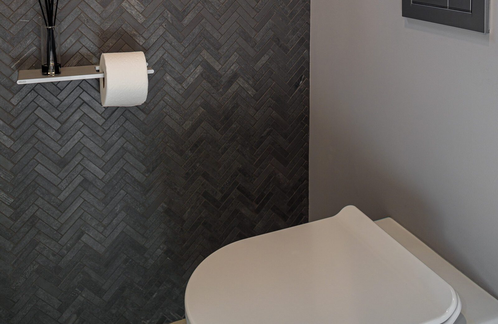 Water closet with dark grey chevron tile wall, white tankless wall mounted toilet